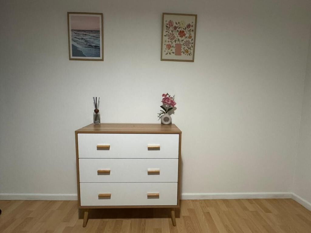 Una habitación con piso de madera, un tocador blanco con tres cajones, un florero con flores y dos cuadros en la pared.
