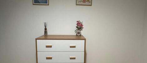 Una habitación con piso de madera, un tocador blanco con tres cajones, un florero con flores y dos cuadros en la pared.