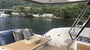 A boat deck with a seating area, a table, and a view of a river with houses and trees. - Sun-Kissed Sailing Adventure in Zadar, Croatia (Bibinje)