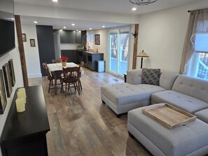 En moderne stue med en grå sofa, et spisebord med stole, et køkken og et stort vindue med udsigt.