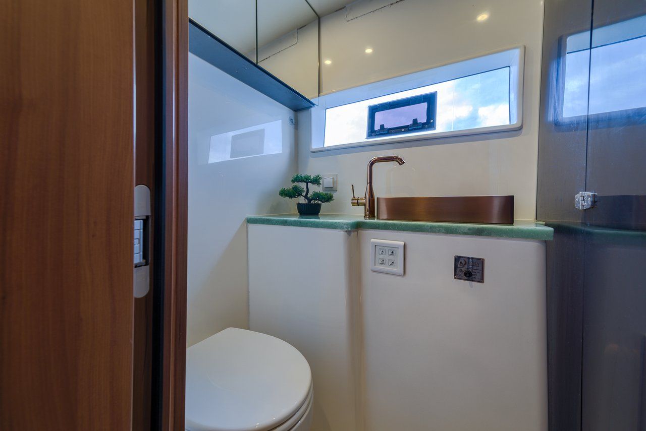A compact bathroom with a white toilet, a small sink with a copper faucet, a potted plant, and a window above the sink.
