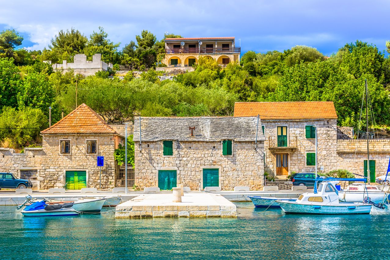 A coastal village with stone buildings, a marina with boats, and a hilltop structure.