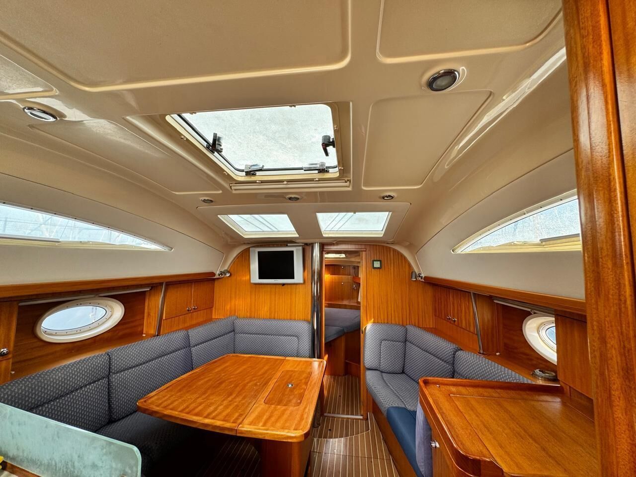 A boat interior with a central dining table, two seating areas with cushioned benches, a TV mounted on the wall, and skylights.