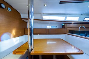Schiffsinnenraum mit Holztisch, weiß gepolsterten Sitzgelegenheiten, Spüle und Fenster.