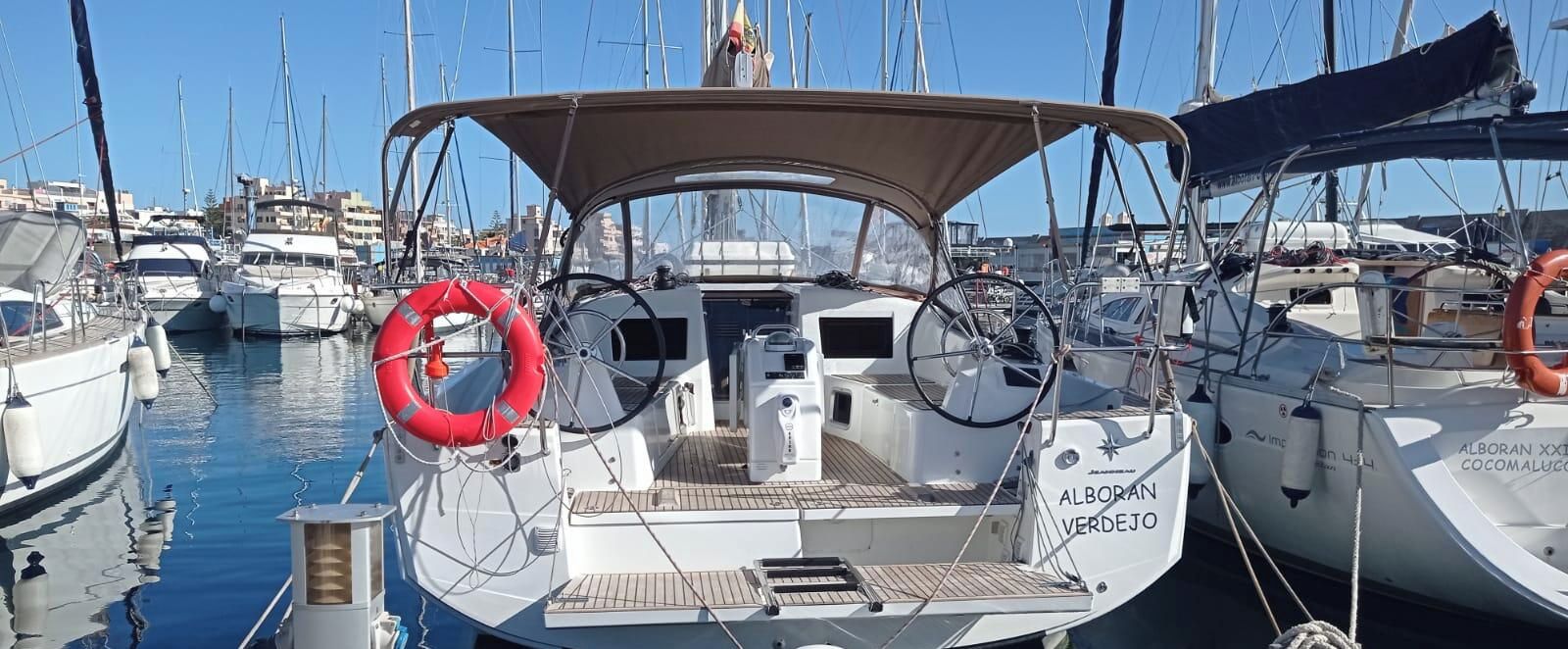 A marina with multiple sailboats docked, featuring lifebuoys, a steering wheel, and a cabin with a name 'ALBORAN VERDEJO'.