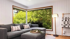 A modern living room with a grey sectional sofa, a floor lamp, and a view of greenery through large windows. - Lake City 5BR Retreat, Hot Tub, 2 Fire Pits, EV, Sleeps 10 (Seattle)