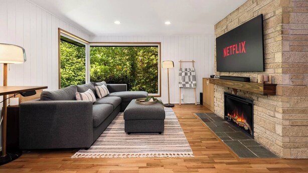 A modern living room with a sectional sofa, a fireplace, a TV mounted on the wall, and a view of greenery outside. - Lake City 5BR Retreat, Hot Tub, 2 Fire Pits, EV, Sleeps 10 (Seattle)
