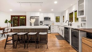 A modern kitchen with a central island, bar stools, stainless steel appliances, and a view of greenery outside. - Lake City 5BR Retreat, Hot Tub, 2 Fire Pits, EV, Sleeps 10 (Seattle)