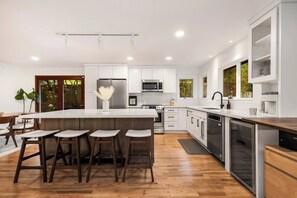 A modern kitchen with a central island, bar stools, stainless steel appliances, and a view of greenery outside. - Lake City 5BR Retreat, Hot Tub, 2 Fire Pits, EV, Sleeps 10 (Seattle)
