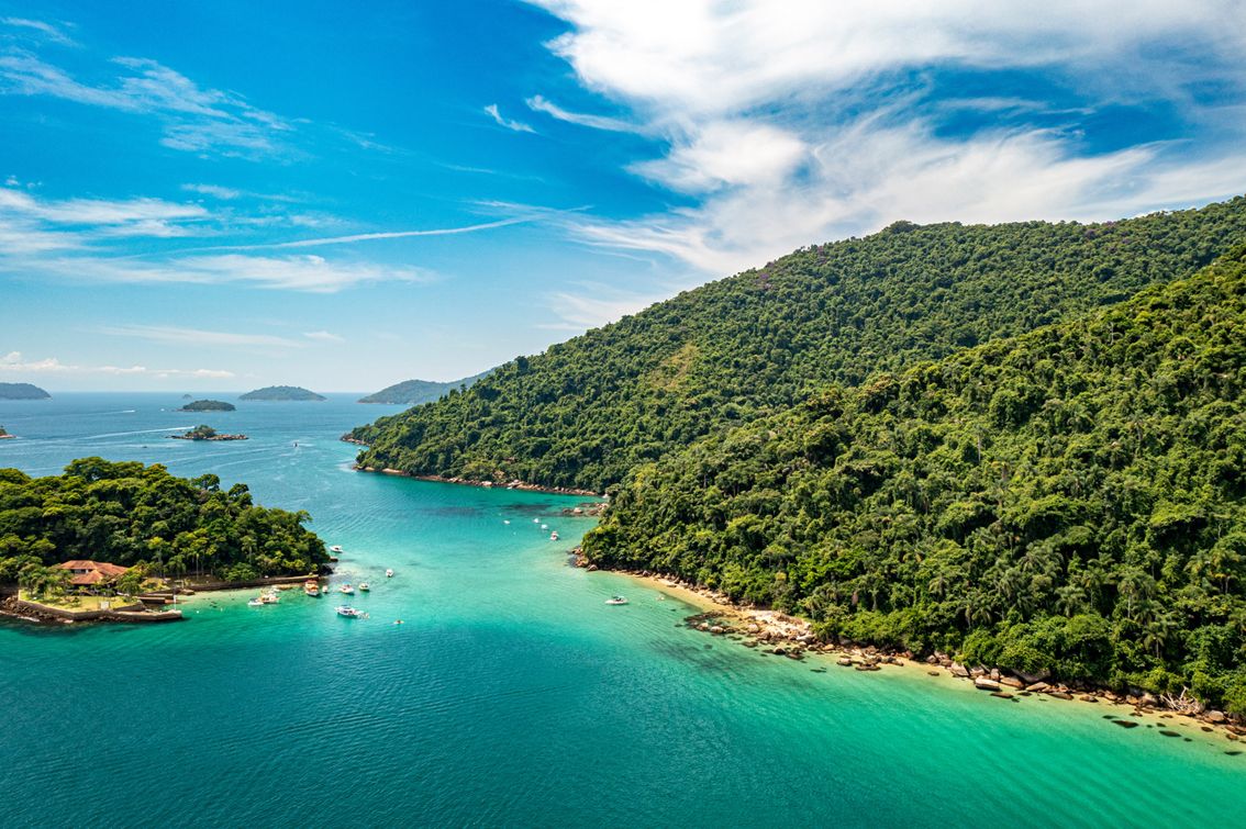 A coastal landscape with a clear view of the sea, a forested hillside, and a small island with boats.