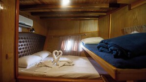Eine Hütte mit zwei Betten, eines mit einer gebügelten Kopfteil und das andere mit schlichtem Kopfteil. Ein Fenster mit Vorhängen ist sichtbar, und eine Klimaanlage ist an der Wand montiert.