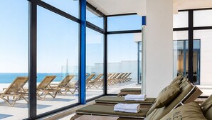 A sunlit room with large windows overlooking a beach, featuring sun loungers with towels and pillows.