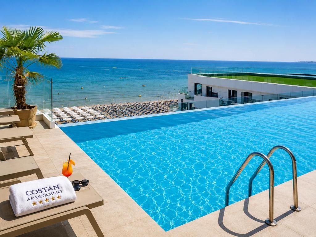 A pool area with a clear view of the ocean, a lounge chair, a towel with 'COSTAN APARTMENT' text, and a palm tree.