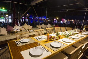 Un espace de restauration en plein air avec une longue table en bois dressée pour un repas, entourée de sièges rembourrés.