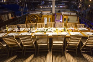Ein Esstisch mit Stühlen ist für eine Mahlzeit gedeckt, mit einem Schiffsrad und Blick auf das Deck.
