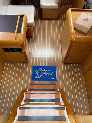 Eine Holztreppe führt zu einem Deckbereich mit einer blauen Matte mit der Aufschrift 'Welcome Aboard' und einem Schild mit Ankersymbol.