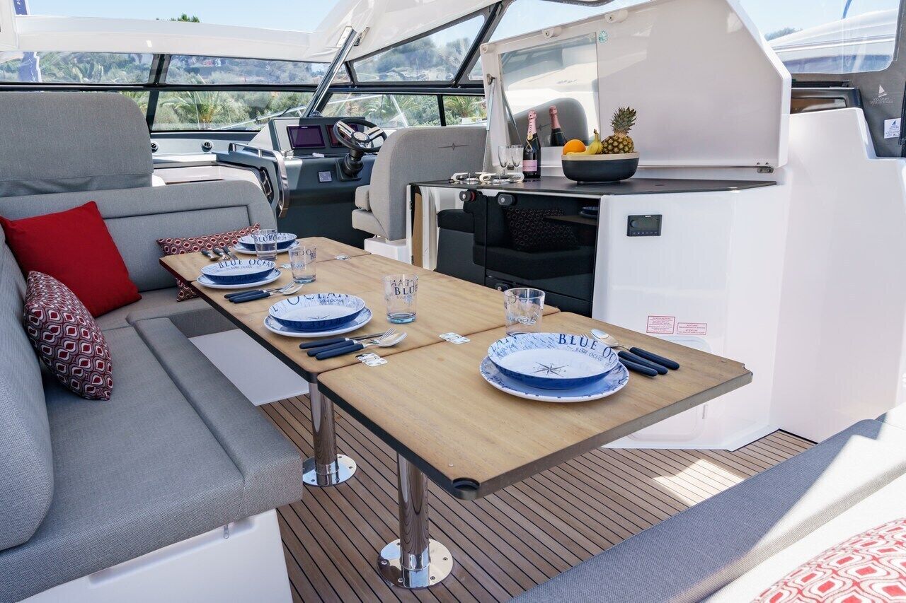 Interior de un barco con comedor, sofá y una mesa para dos personas, preparada con platos y cubiertos.