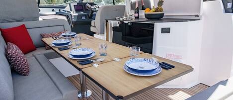 Interior de un barco con comedor, sofá y una mesa para dos personas, preparada con platos y cubiertos.
