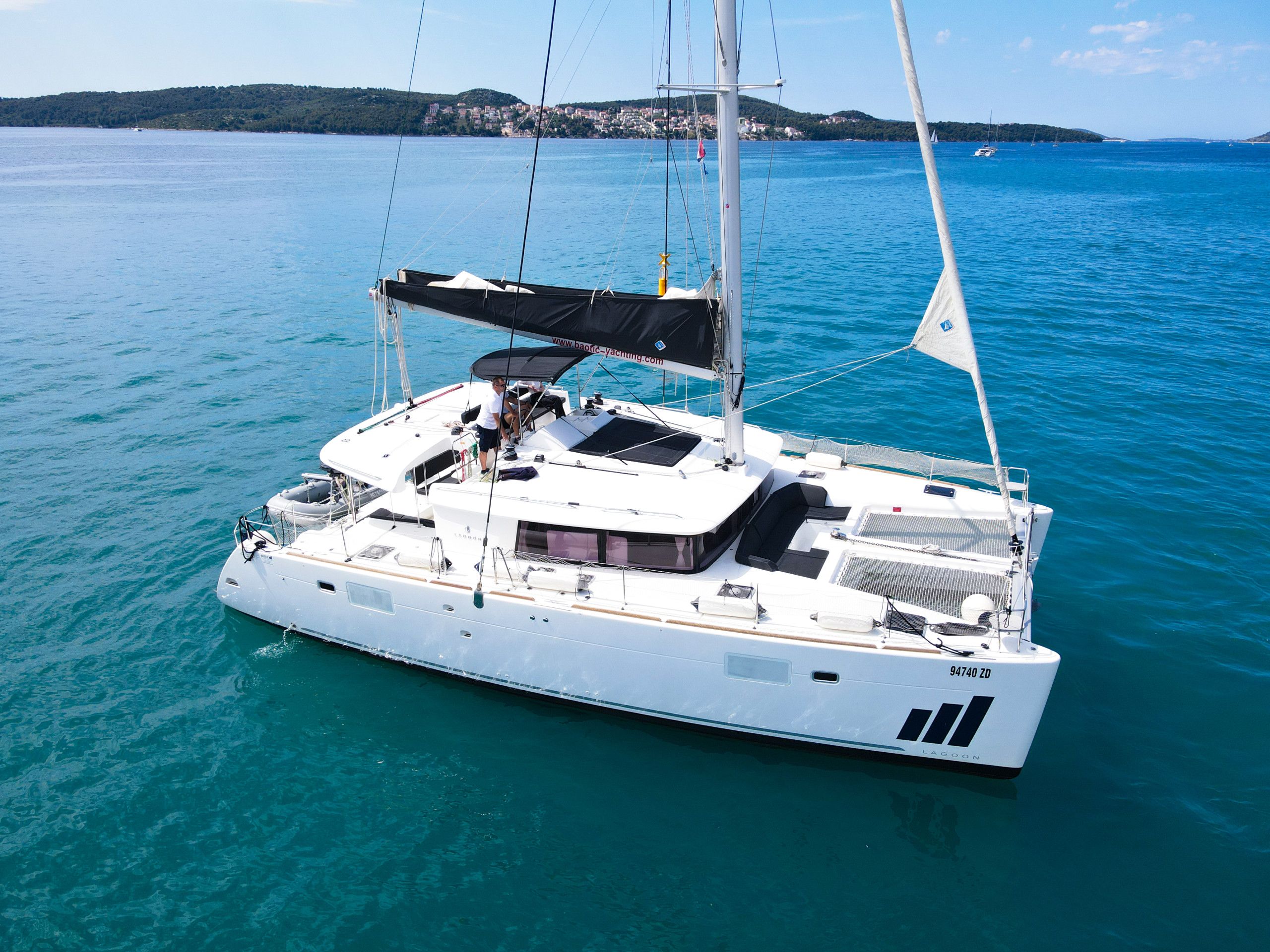 Un velero blanco con cabina y asientos en la cubierta, fondeado en aguas azules tranquilas, con un pueblo costero al fondo.