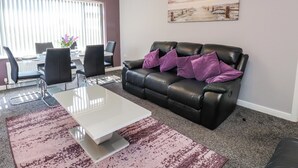 A living room with a black leather sofa, a white coffee table, and a dining area with a table and chairs. - Ger y Mynydd, 2 Cae Ty Coch (Brynteg)