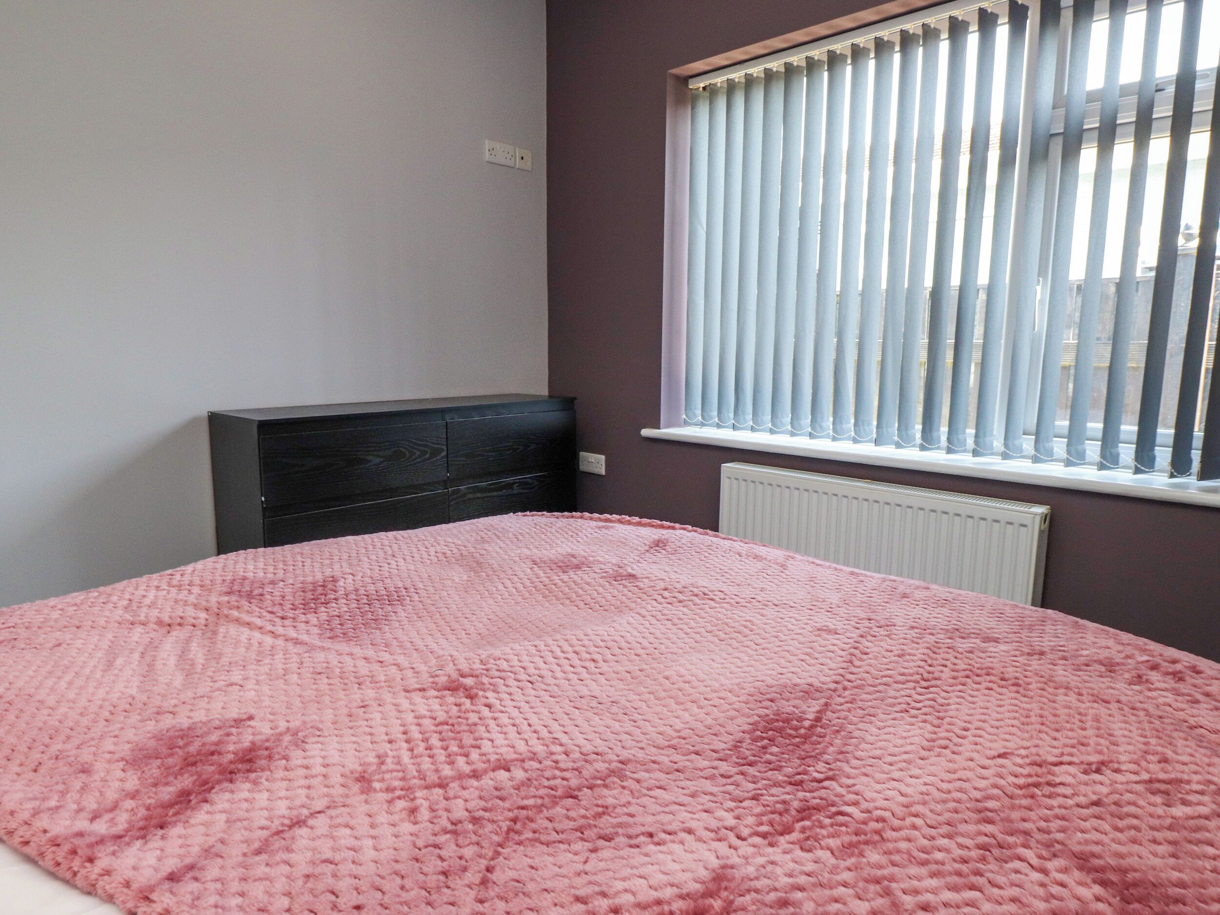 A bedroom with a bed, a dark headboard, a window with blinds, and a radiator.