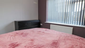 A bedroom with a bed, a dark headboard, a window with blinds, and a radiator.