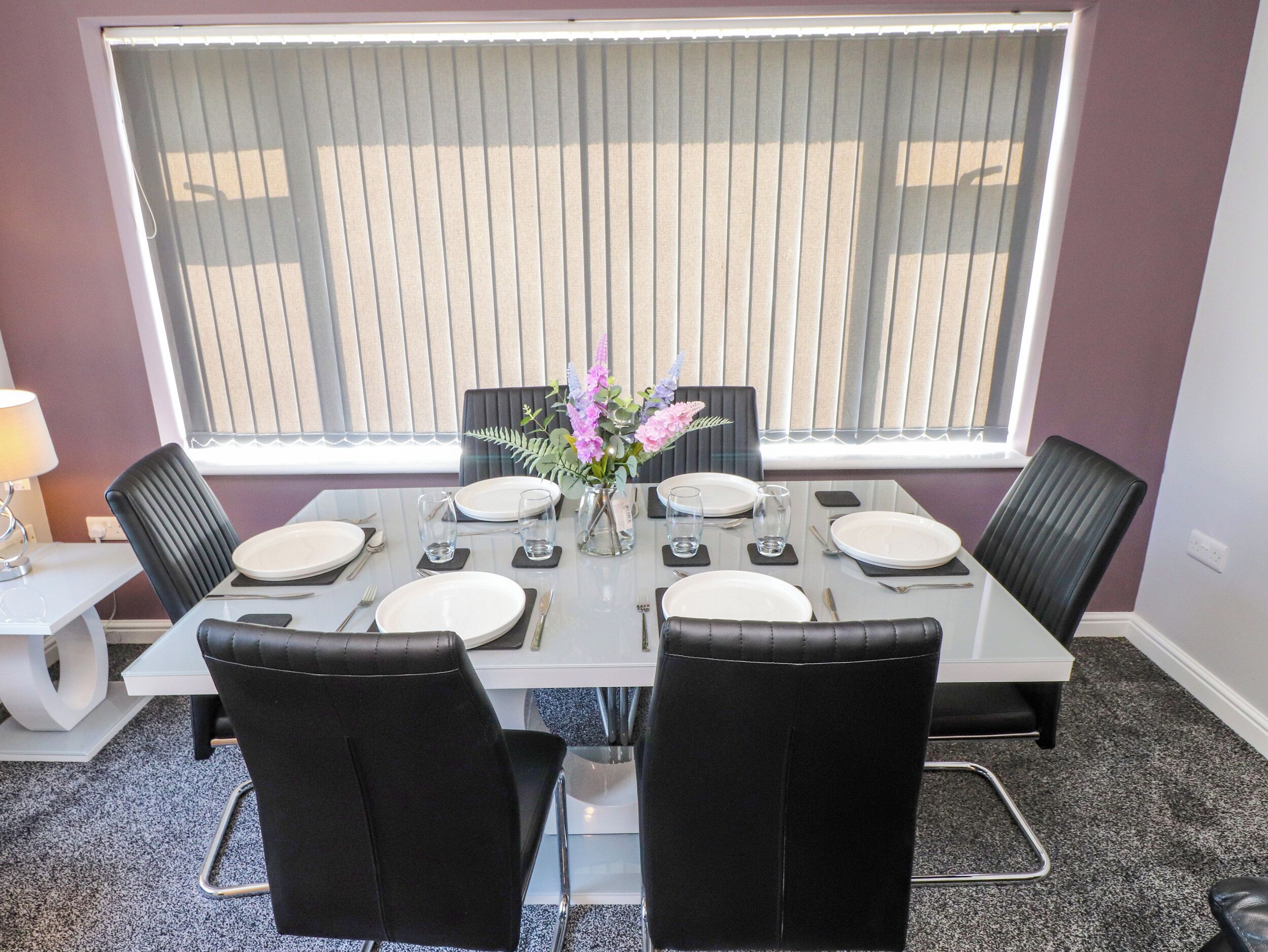 A dining area with a table set for four, chairs, a vase with flowers, and a window with blinds.