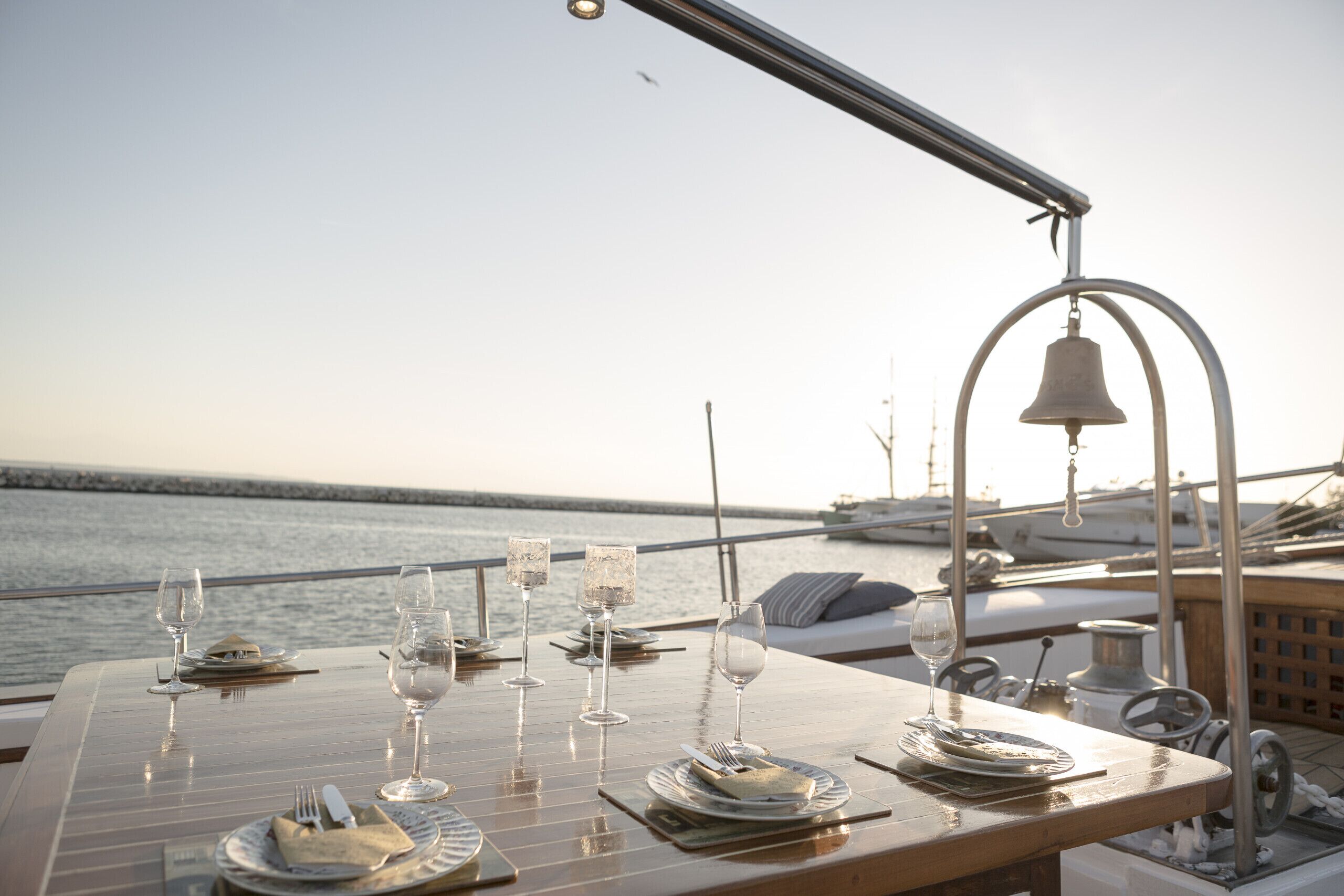 A table set for a meal with glasses and plates, a bell, and a view of the water.