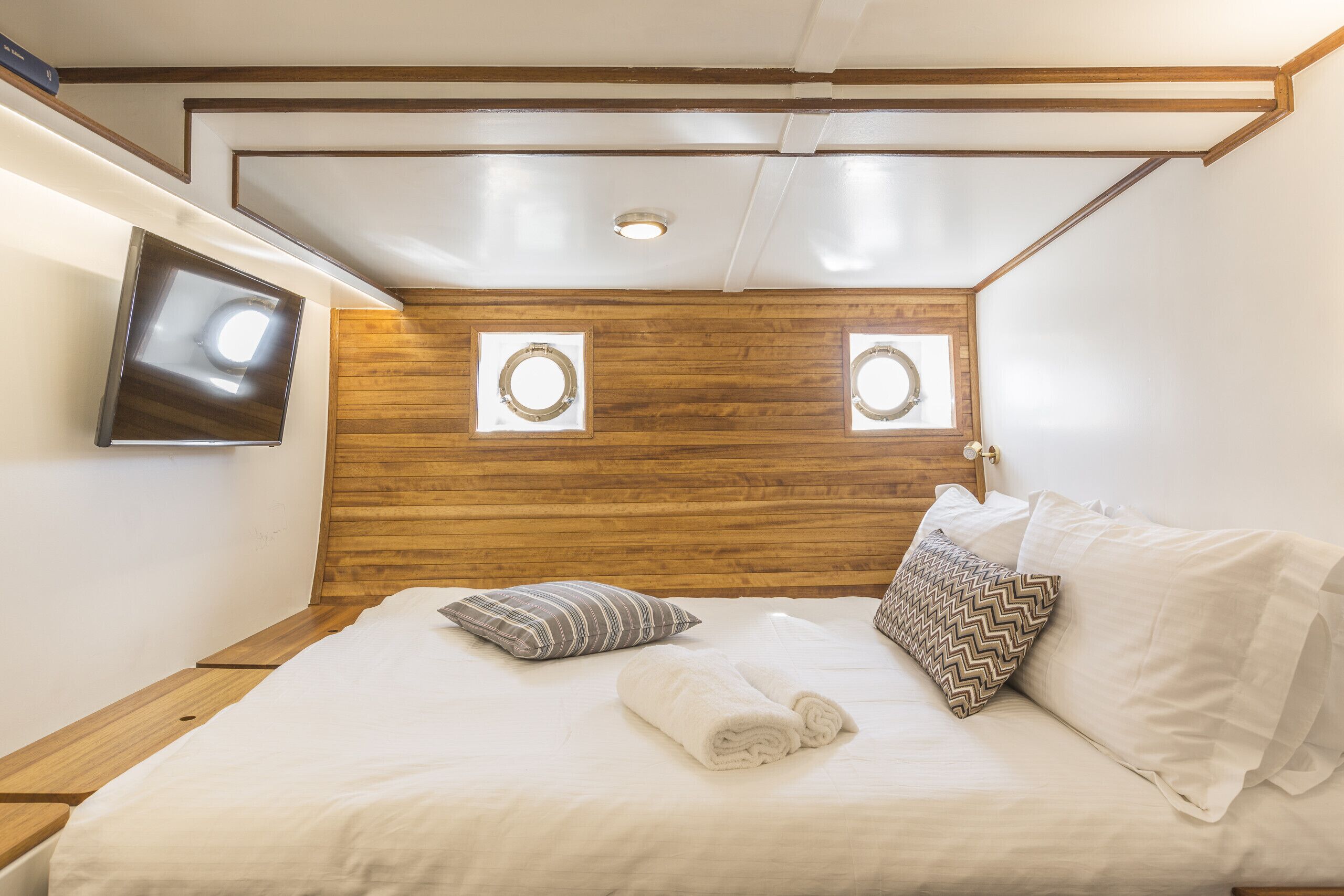 A hotel room with a wooden headboard, two wall-mounted circular light fixtures, a flat-screen TV, and a neatly made bed with white linens and patterned pillows.