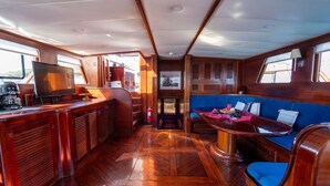 A boat interior with a wooden floor, a seating area with blue cushions, a table with a red object, and a television on a stand. - Sun-Kissed Sailing Adventure in Islas Sarónicas (Alimos)