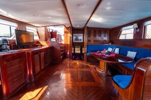 A boat interior with a wooden floor, a seating area with blue cushions, a table with a red object, and a television on a stand.