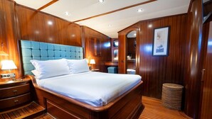 A wooden-paneled bedroom with a tufted headboard, a bed with white linens, a nightstand with a lamp, and a small waste bin. - Sun-Kissed Sailing Adventure in Islas Sarónicas (Alimos)