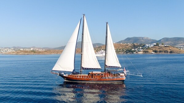A boat with two masts and sails on a body of water with a hilly landscape in the background.