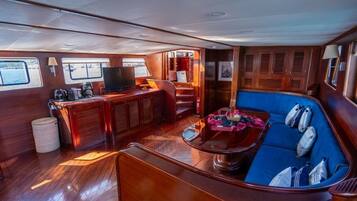 Interior de un barco con cubierta de madera, zona de estar con cojines, estación de café y televisor.