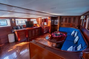 A boat interior with a wooden deck, a seating area with cushions, a coffee station, and a television.