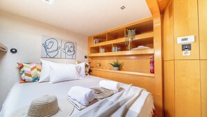 A bedroom with a bed, a wooden wardrobe, a shelf with books and a plant, and a ceiling light.