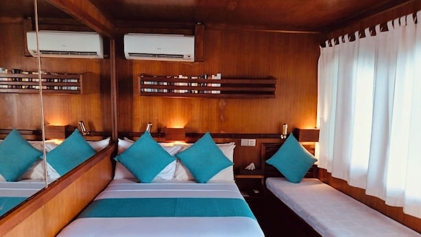 Una cabaña con dos camas, cada una con cabecero de madera, ropa de cama blanca y almohadas color turquesa. Hay unidades de aire acondicionado sobre las camas y una ventana con cortinas blancas a la derecha.