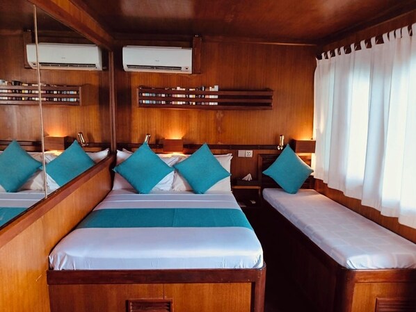 Une cabine équipée de deux lits, chacun doté d’une tête de lit en bois, de draps blancs et d’oreillers turquoise. Des unités de climatisation sont installées au-dessus des lits, et une fenêtre avec des rideaux blancs se trouve sur le côté droit.