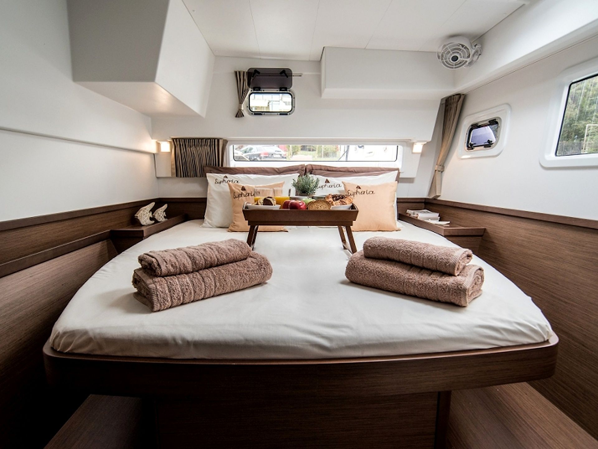 A boat cabin with a bed, a small table with a tray of food, and a window with curtains.