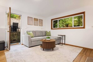 A living room with a grey sofa, a round wooden coffee table, and a large window with a view of trees. - Lake City Downstairs 2BR • Movie Nights + Fireplace (Seattle)