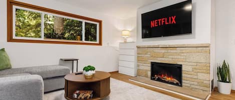 Una sala de estar con chimenea, un televisor montado encima y un ventanal con vistas a árboles.