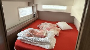 En kompakt seng med rød madras og flere puder, et lille vindue med persienner samt en ventil i loftet.