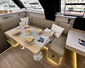 Ein Yacht-Interieur mit Essbereich, inklusive Tisch für vier Personen, gepolsterten Sitzgelegenheiten und einem Fenster mit Blick auf eine Marina.