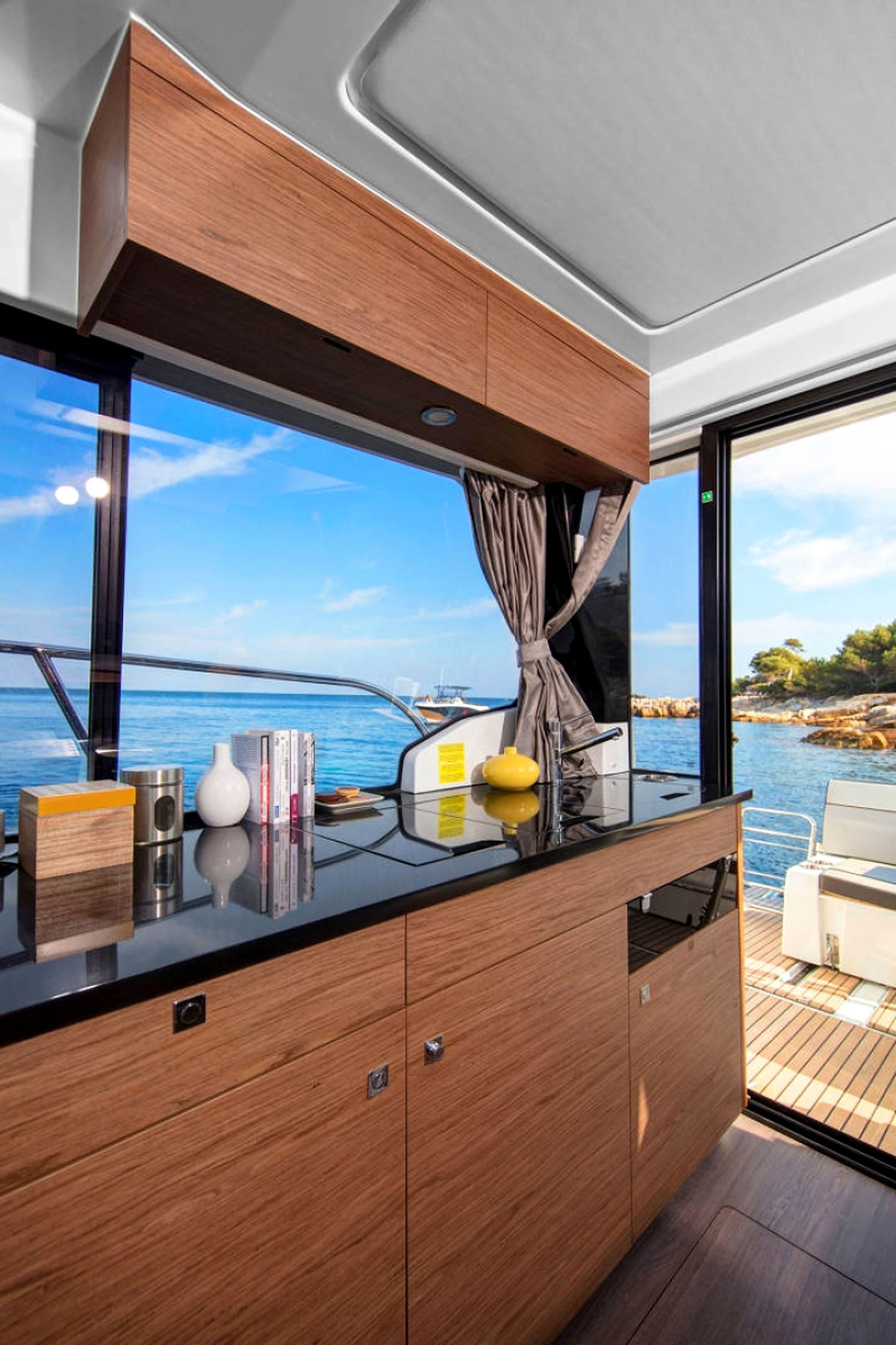 A modern kitchen with wooden cabinets, a black countertop, and a view of the sea through large windows.