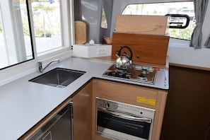A compact kitchenette with a sink, stove, and oven, wooden cabinetry, and a window with a view of the outside. - Dubrovnik Delight: A Sun-Kissed Sailing Experience (Komolac)