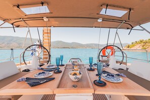A boat's dining area with a table set for two, a steering wheel, and a view of the sea and hills. - Sun-Kissed Sailing Adventure in Islas Sarónicas (Alimos)