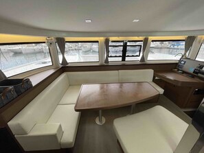 A boat interior with a central seating area, a control panel, and windows providing views of the water.