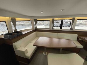 Interior de barco con zona de estar central, ventanas con cortinas y vista al puerto deportivo.