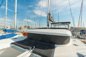 Cubierta de barco con zona de asientos acolchada, un mástil y otros barcos al fondo.