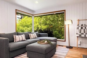 A modern living room with a grey sectional sofa, a floor lamp, and a view of greenery through large windows. - Lake City 3BR Retreat with Hot Tub, Fire Pit & EV (Seattle)
